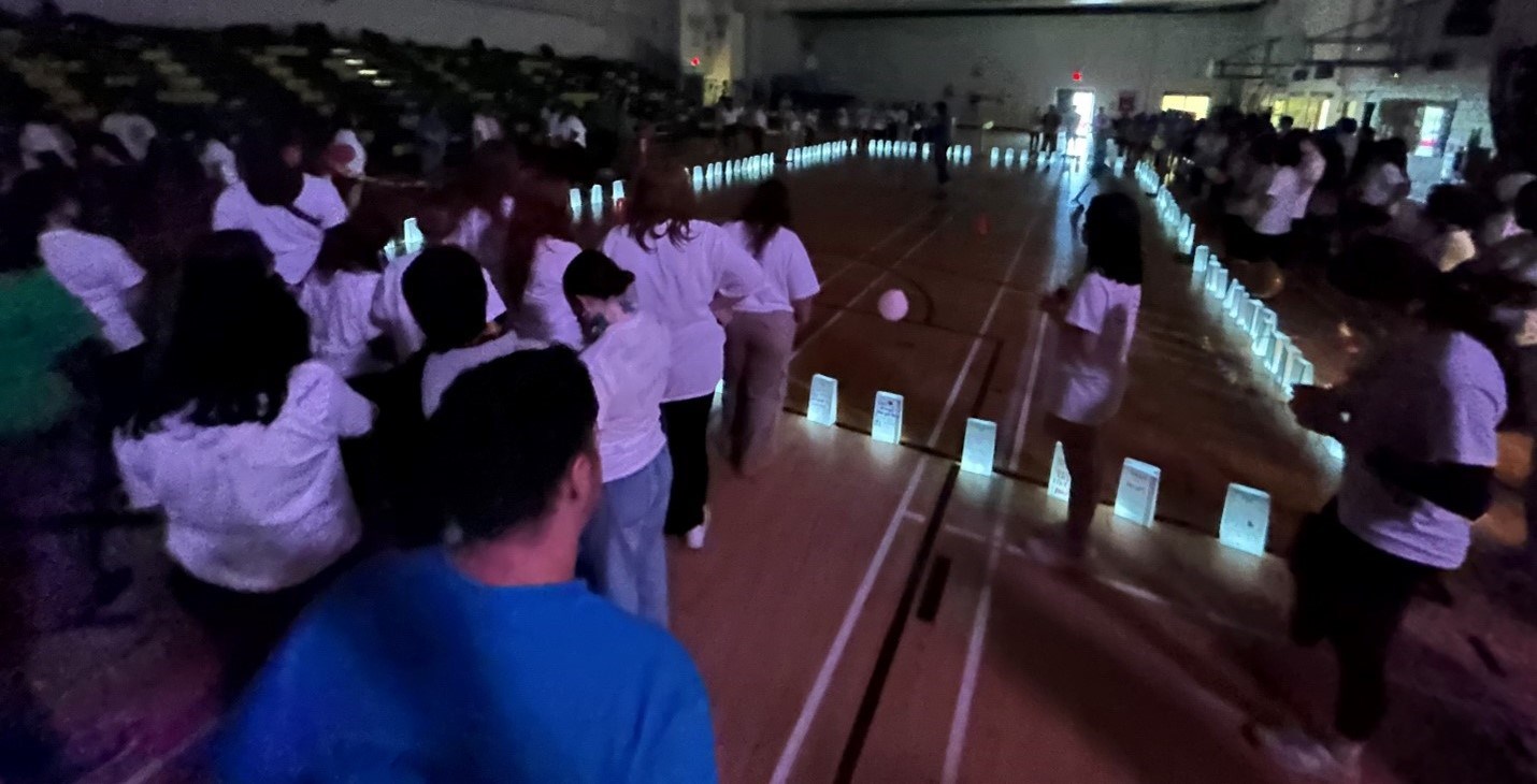 Students and staff in a dark gym huddle around rows of illuminated bags with messages written on them to honour those who have passed away from cancer, are battling cancer, or have survived cancer.