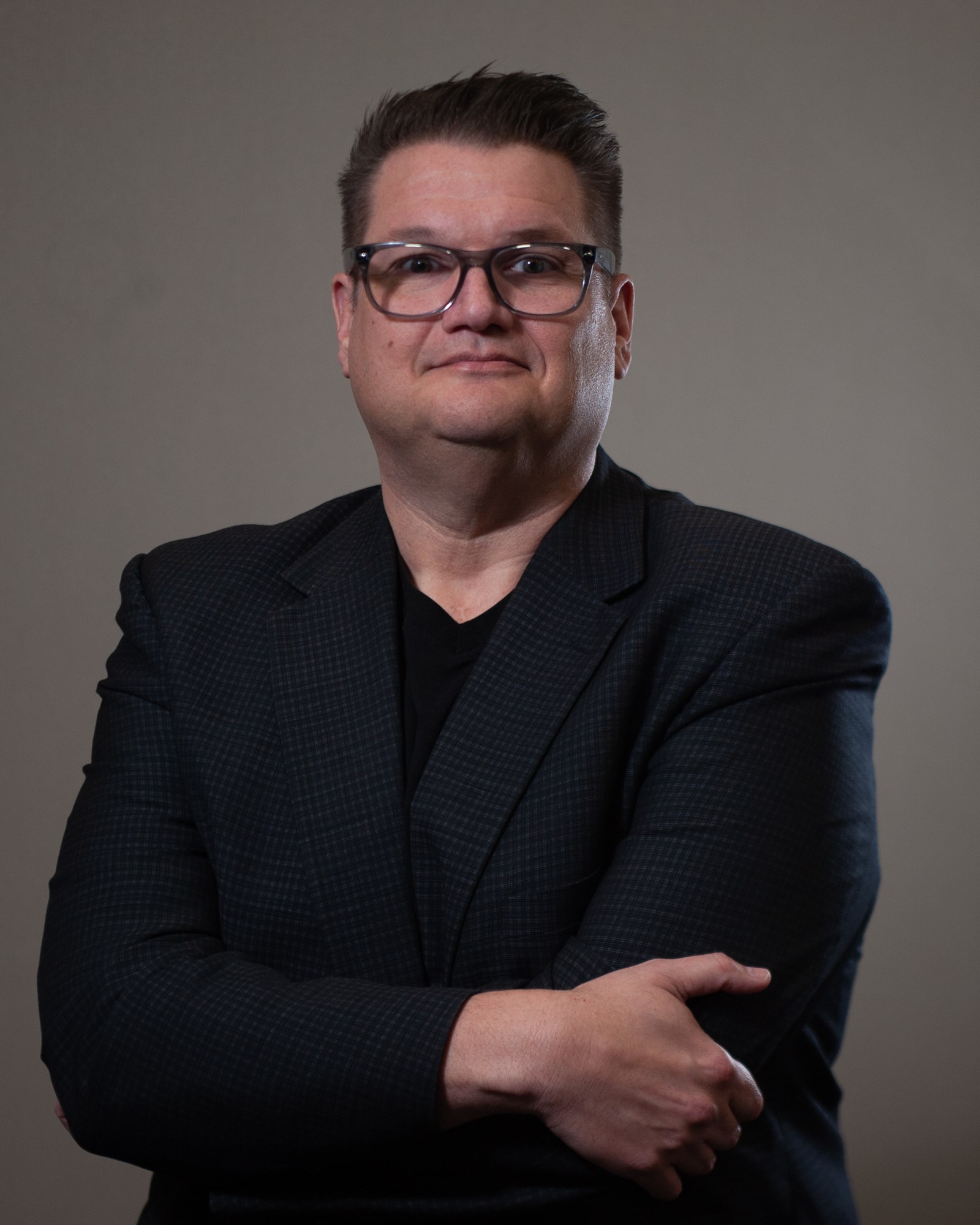 Corey Kapilik in a black suit jacket and t-shirt with one arm crossed over his chest holding his elbow against a grey background.