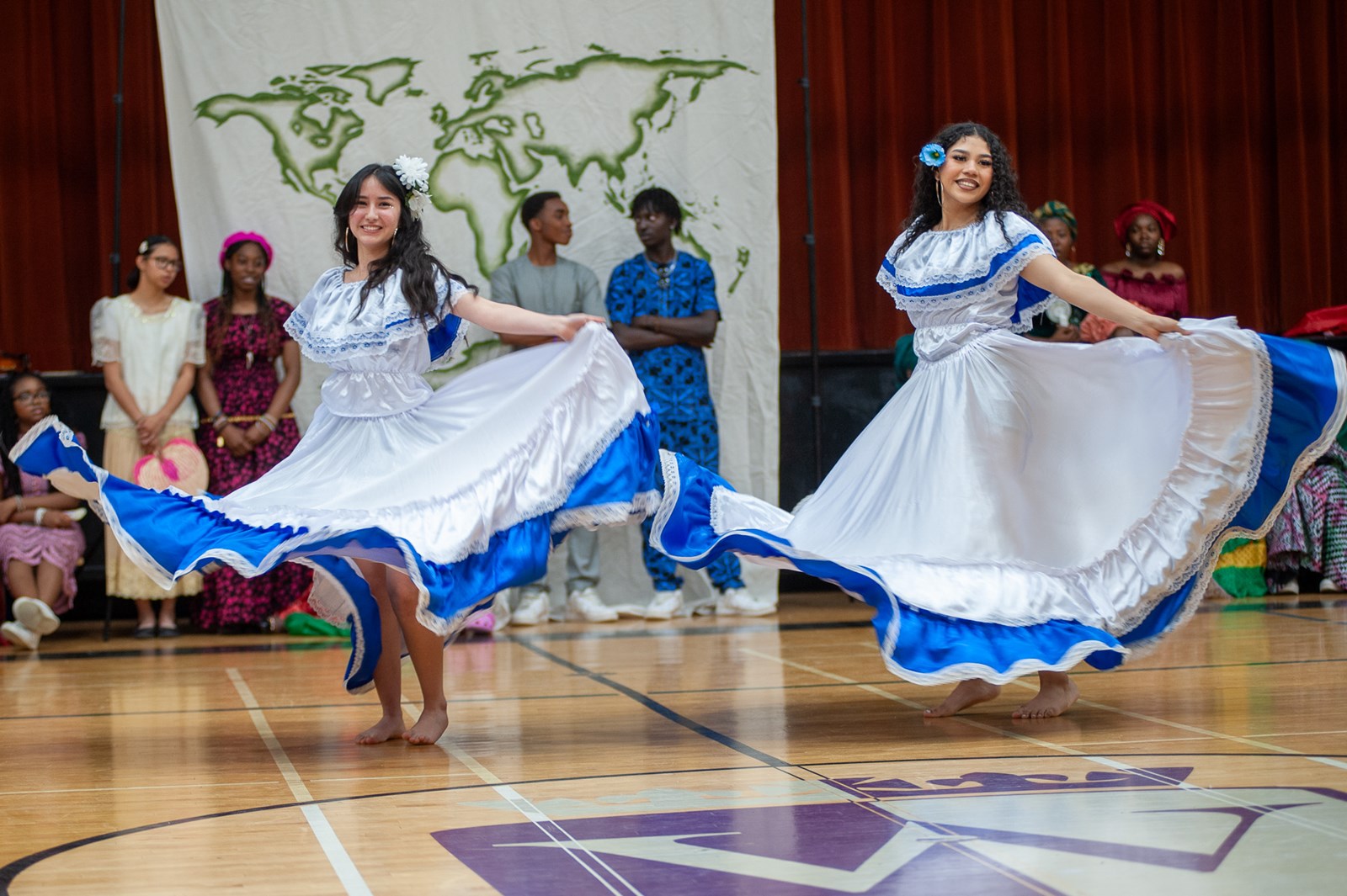 Two female students perform a Columbian dance wearing blue and white dresses that twirl through the air as they spin.