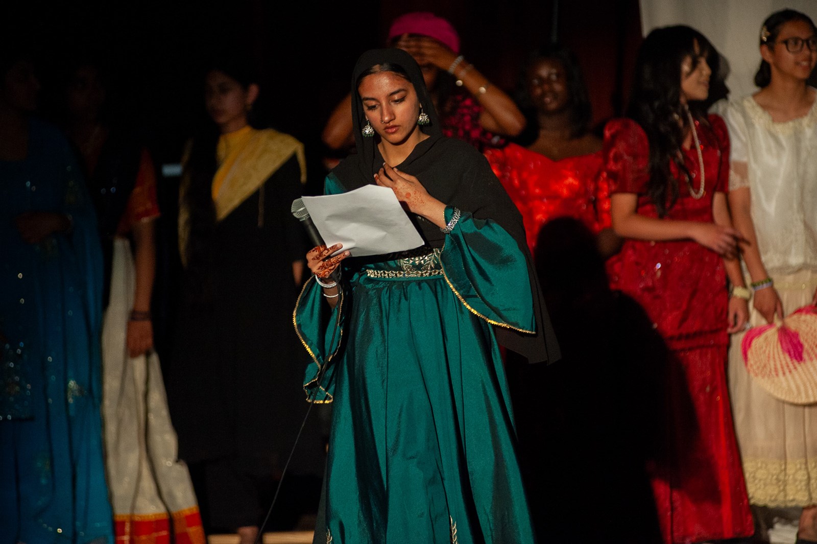 Cultural Gala M.C. Shafia Razzaq reads a piece of paper and holds a microphone. There are students in a variety cultural clothing in the shadows behind her.