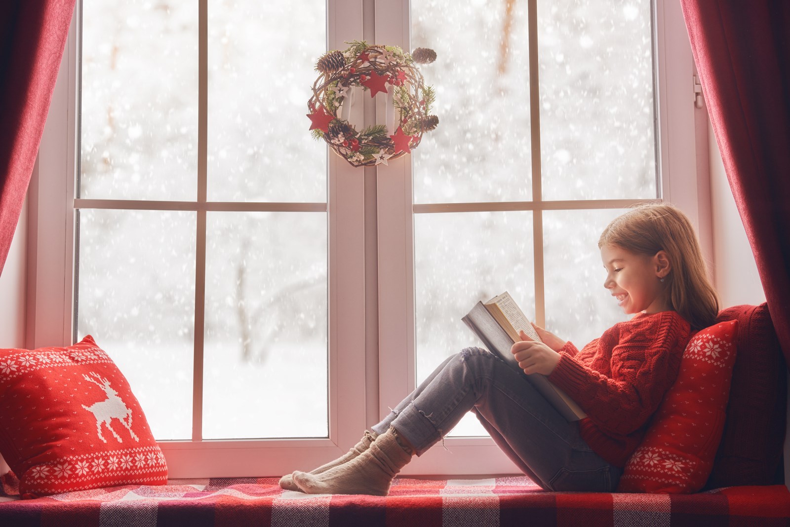 Cute little girl sitting by the snowy window and reading a book.